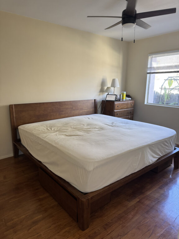A tidy bedroom with a wooden bed frame, white mattress, wooden dresser, lamp, and a ceiling fan, with sunlight coming through a window.