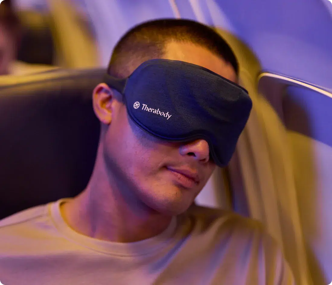 A man wearing a dark eye mask labeled "Therabody" rests on an airplane seat under soft, blueish lighting.