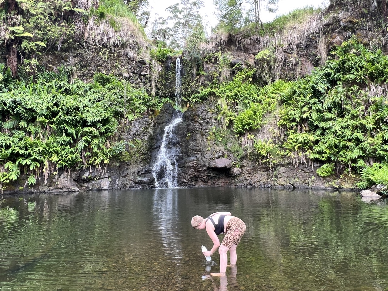A person bends down to touch the water in a shallow pool beneath a small, thin waterfall surrounded by lush greenery.