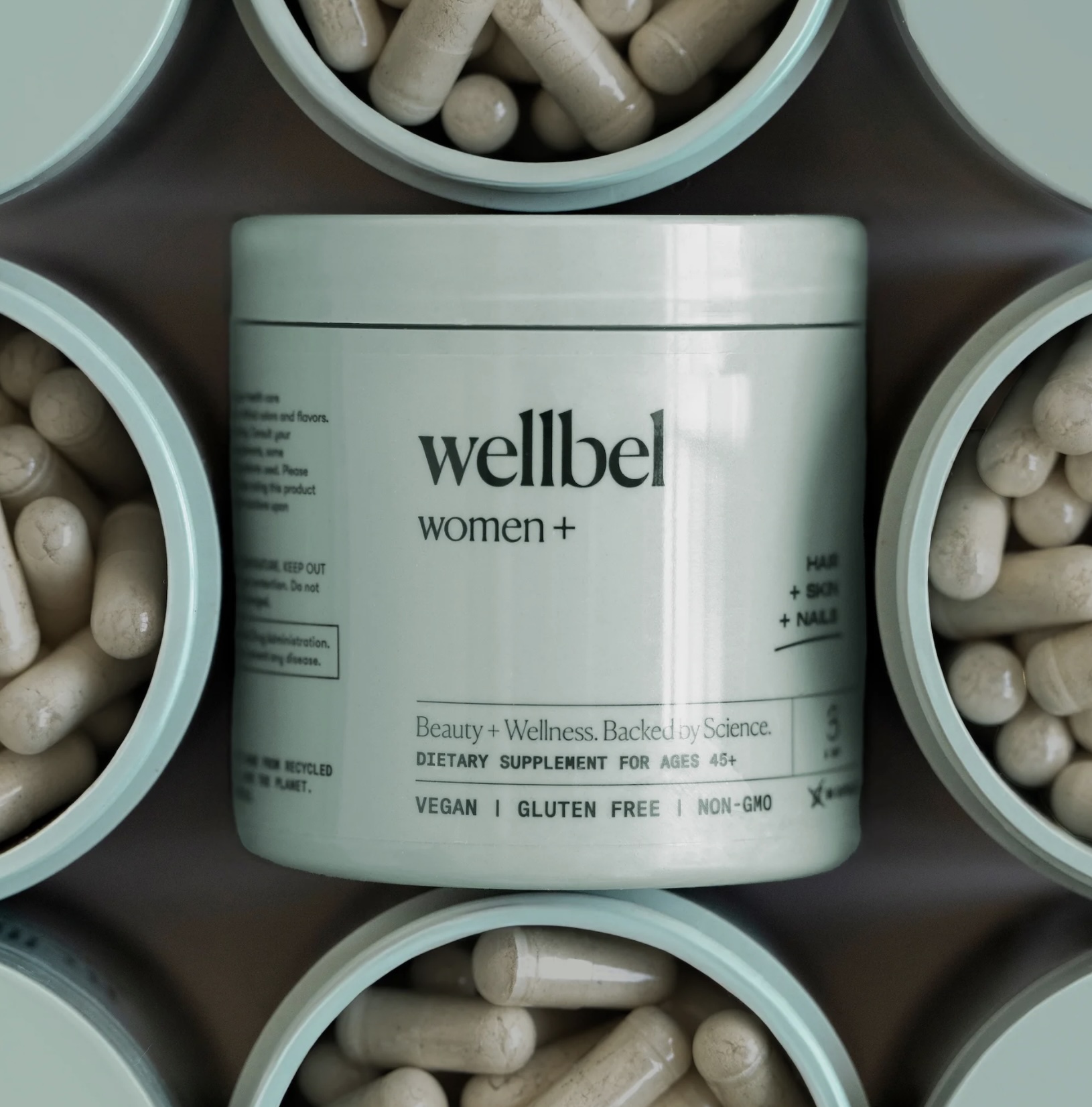 A wellbel women+ dietary supplement container is surrounded by open jars filled with capsules. The label highlights hair, skin, and nail benefits, and notes the product is vegan, gluten-free, and non-GMO.