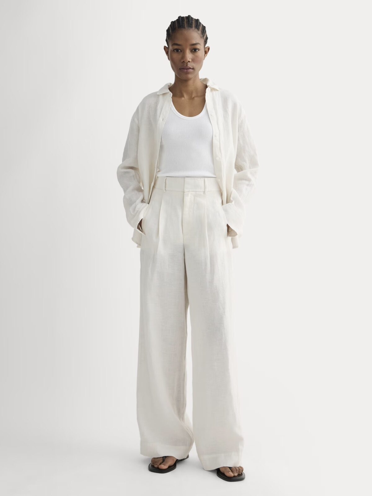 A person stands facing forward wearing a white tank top, a cream-colored button-up shirt, wide-leg trousers, and black sandals.