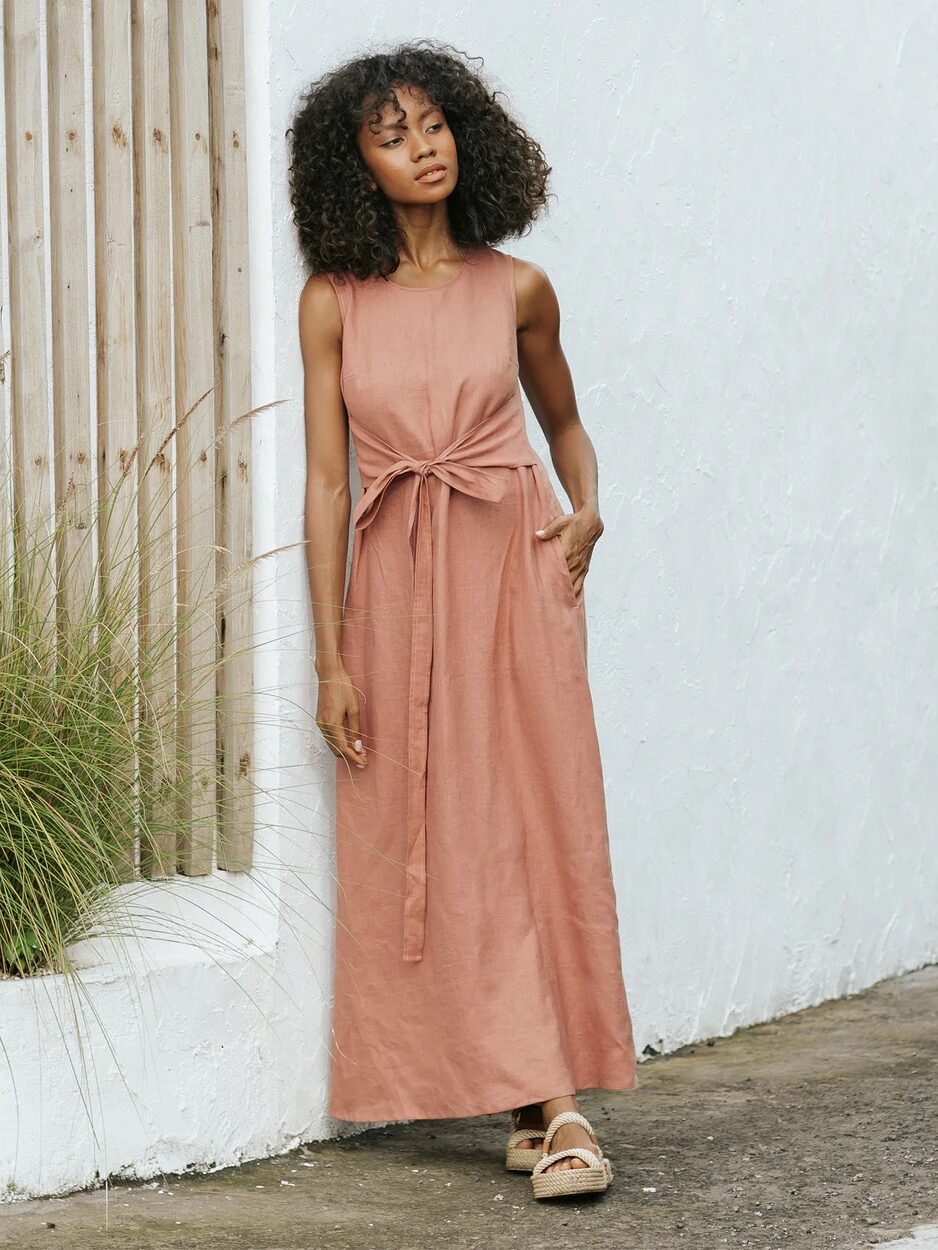 A woman stands outdoors wearing a sleeveless, ankle-length, light brown dress with a front tie and sandals, next to a white wall and wooden fence.