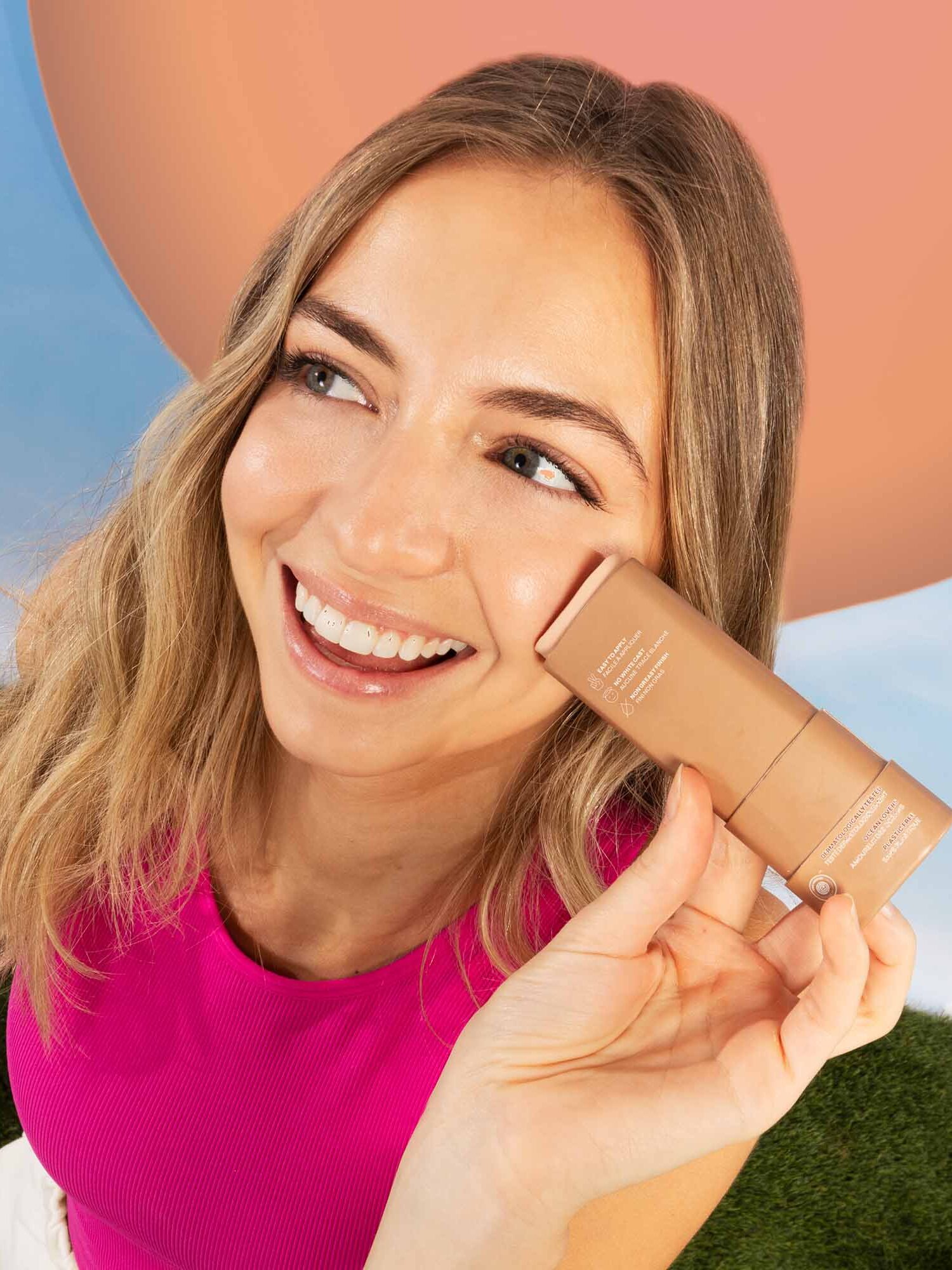 A smiling woman in a pink top holds a tan-colored cosmetic product next to her face outdoors with a blue sky and large peach circle in the background.