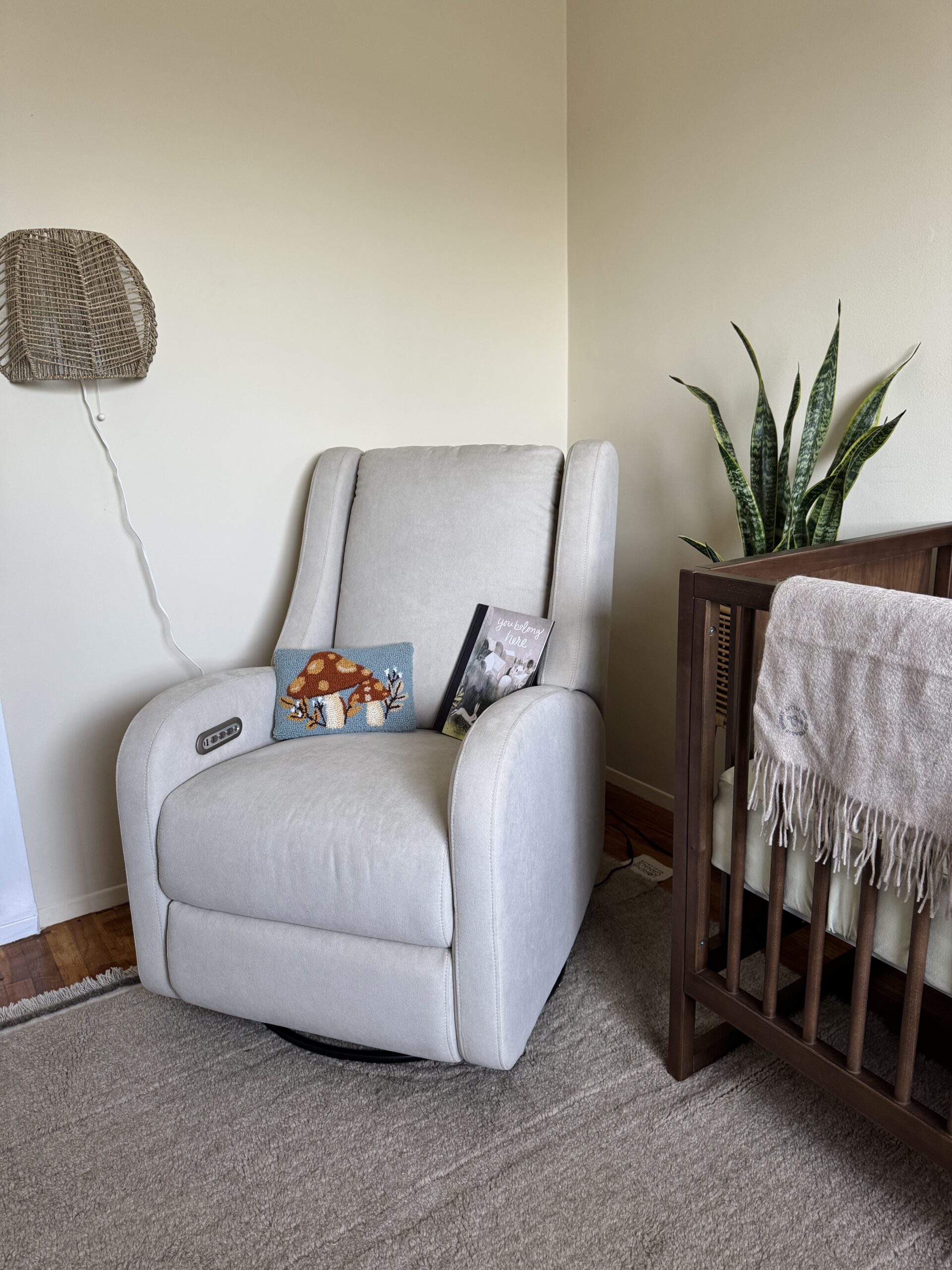 A beige recliner with two children's books on the seat is positioned next to a wooden crib, a potted snake plant, and a wall-mounted rattan lamp in a nursery.