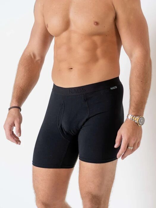 A shirtless man wearing black boxer briefs stands against a plain white background, with a watch on his left wrist.