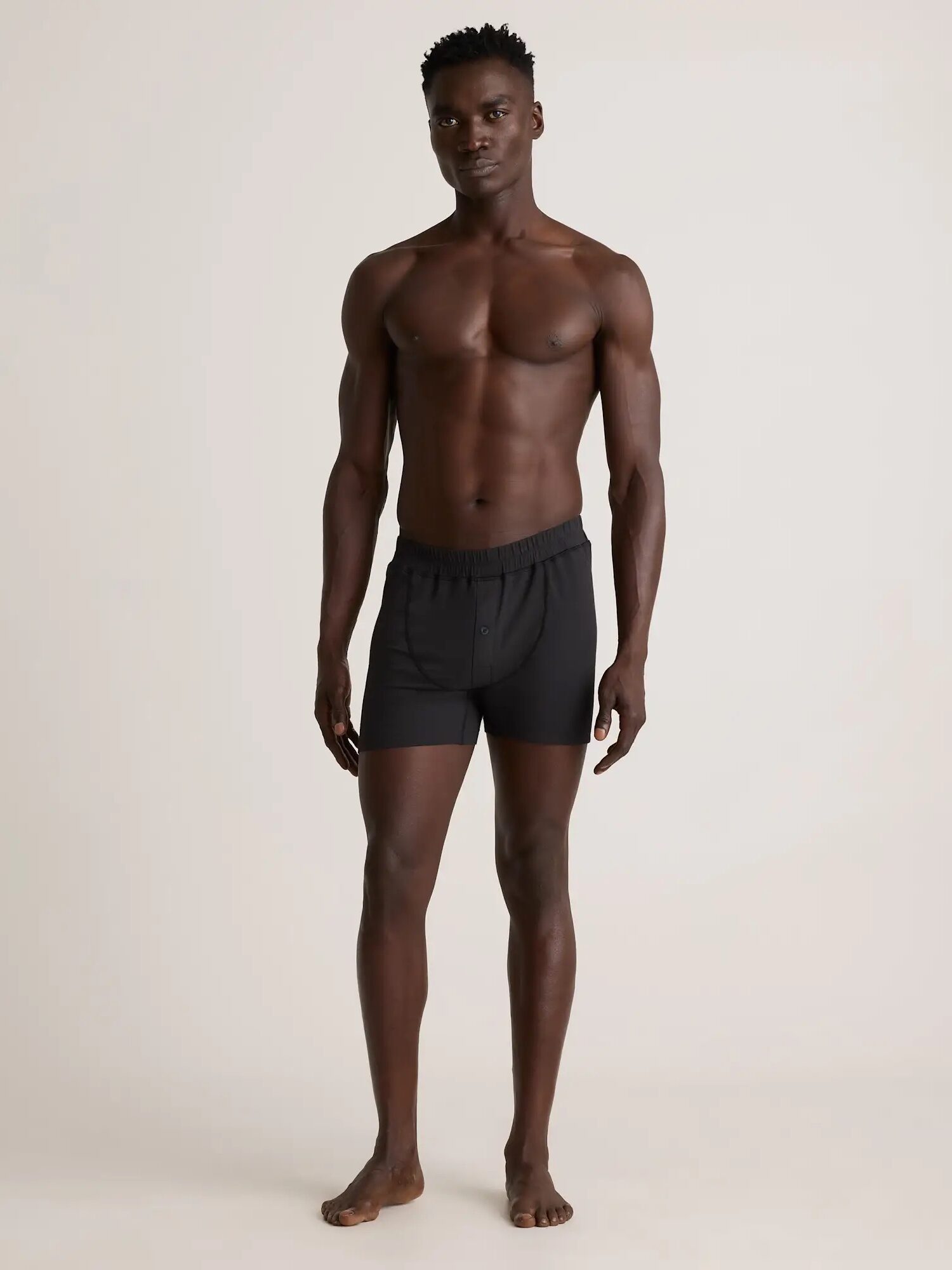 A man stands barefoot against a plain background, wearing black boxer shorts and no shirt.