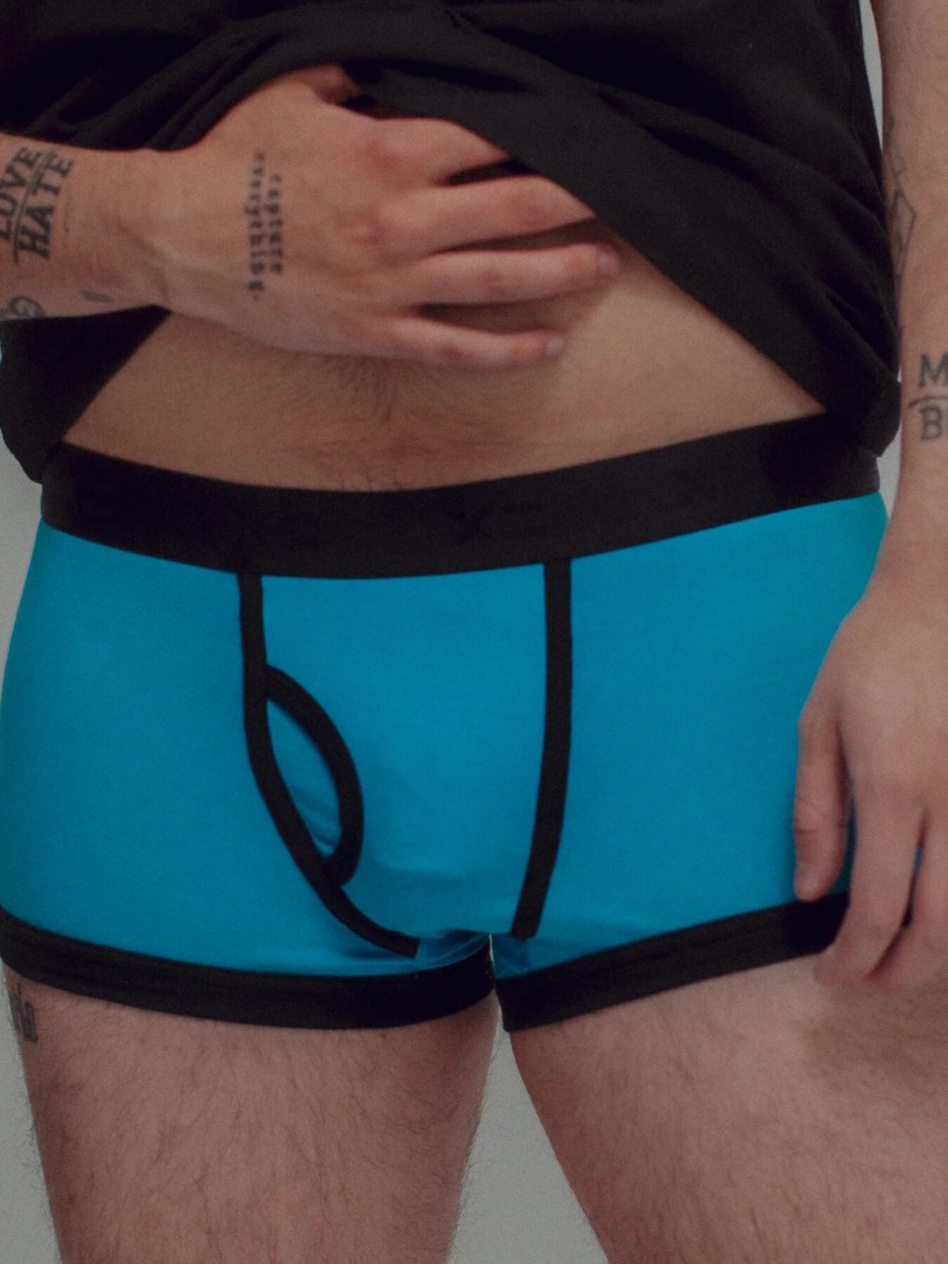A person lifts their shirt to reveal bright blue boxer briefs with black trim; tattoos are visible on their arms and hands.