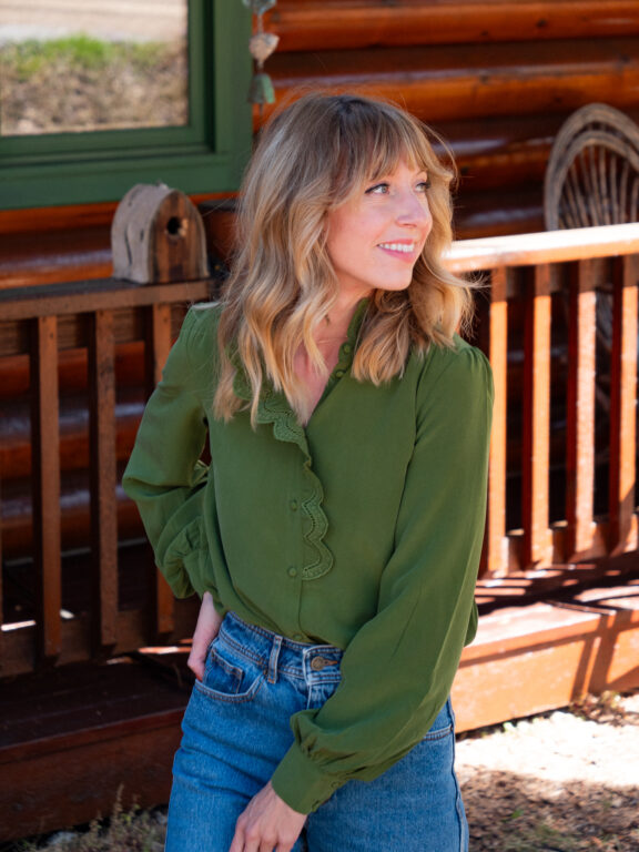 Woman with wavy blonde hair wearing a green long-sleeve blouse and blue jeans stands outside in front of a wooden cabin, looking to her left and smiling, effortlessly channeling french girl fashion.