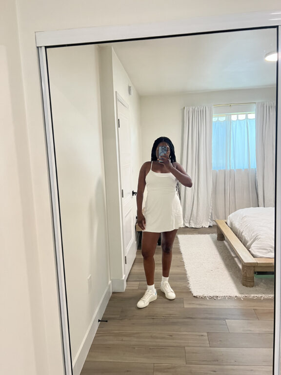 A person wearing a white dress and sneakers takes a mirror selfie in a bedroom with white walls, curtains, and bedding.