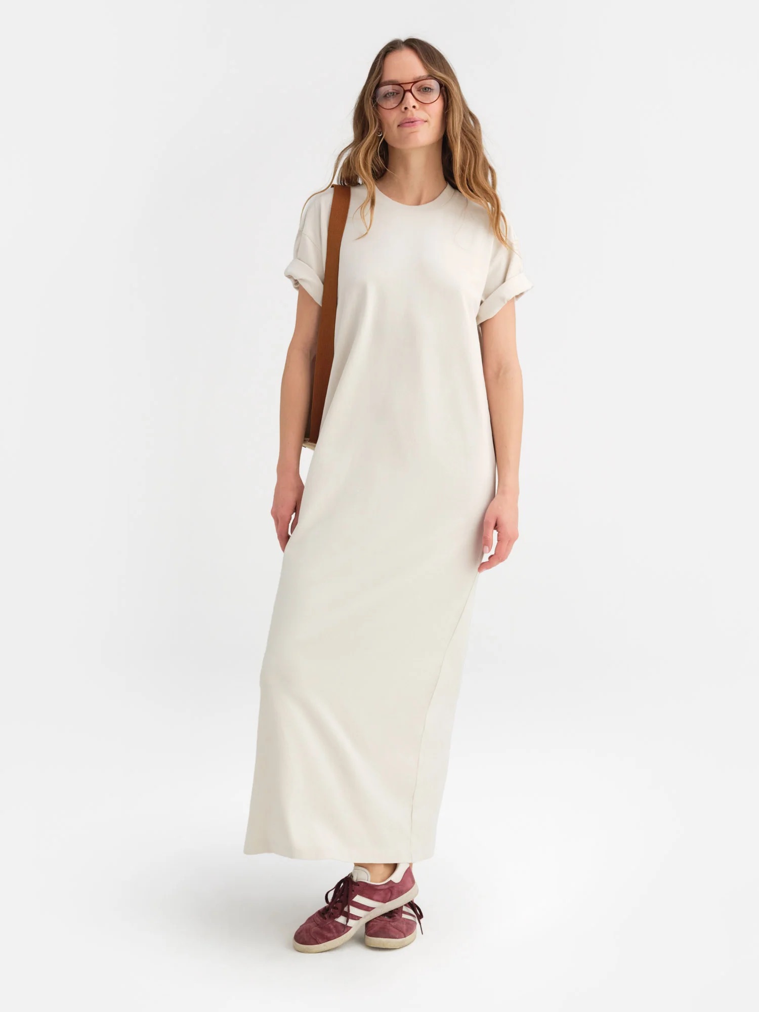 A woman wearing glasses, a loose-fitting cream-colored maxi dress, and red sneakers stands against a plain white background.