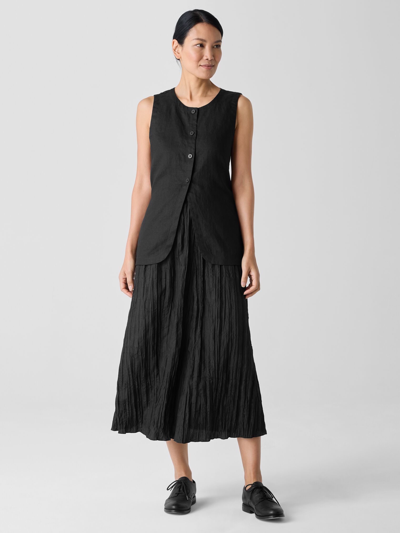 A woman stands against a plain background wearing a sleeveless black buttoned top and a black ankle-length pleated skirt, paired with black shoes.