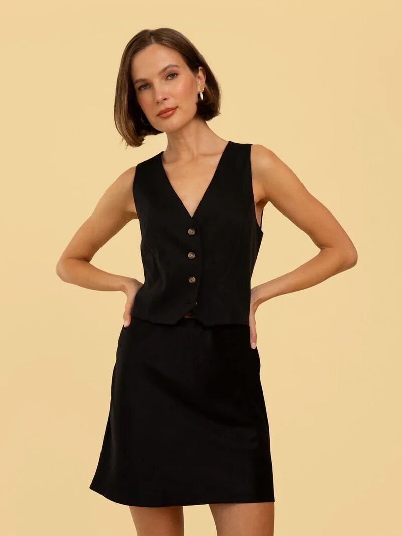 A woman wearing a sleeveless black buttoned vest and matching black skirt stands against a plain beige background.