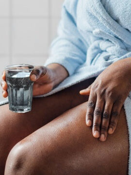 A person in a light blue bathrobe sits with one hand holding a glass of water and the other hand resting on their thigh.