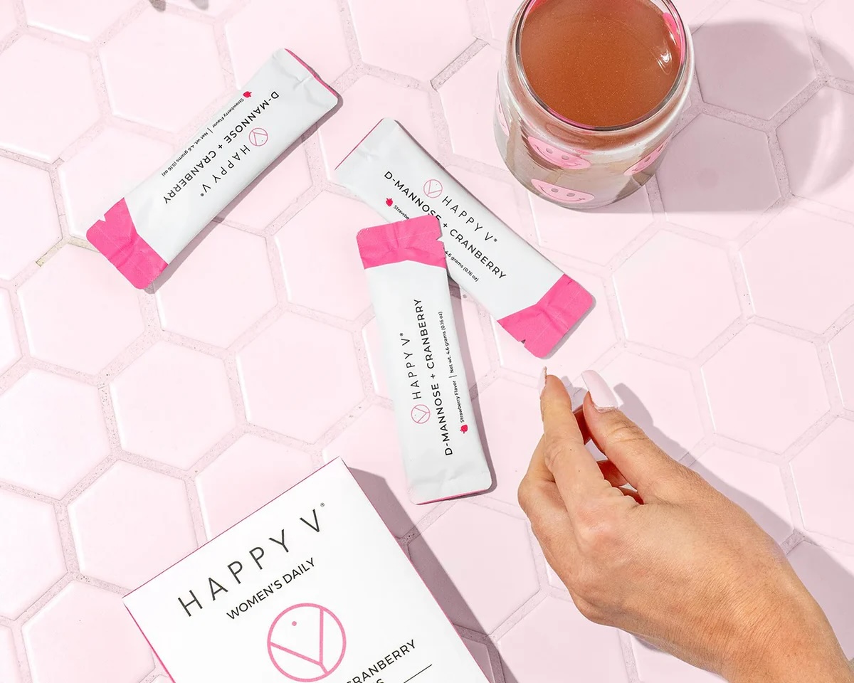 A hand reaches for a pink packet labeled "Happy V D-Mannose + Cranberry" on a pink hexagonal tile surface with a glass of pink liquid and other supplement packets nearby.
