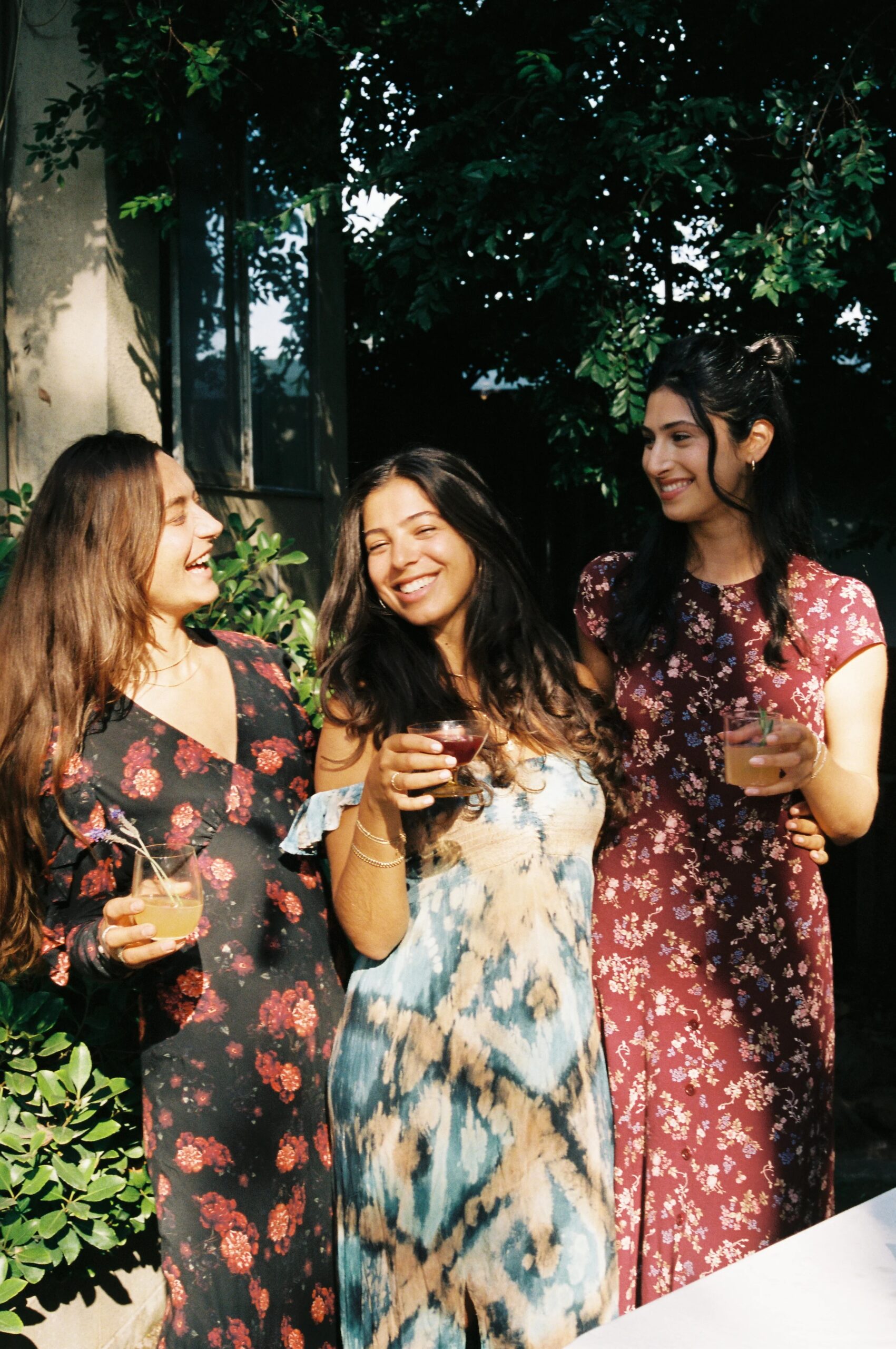 Three women in patterned dresses stand outdoors holding drinks, smiling and talking together in natural sunlight with greenery in the background.