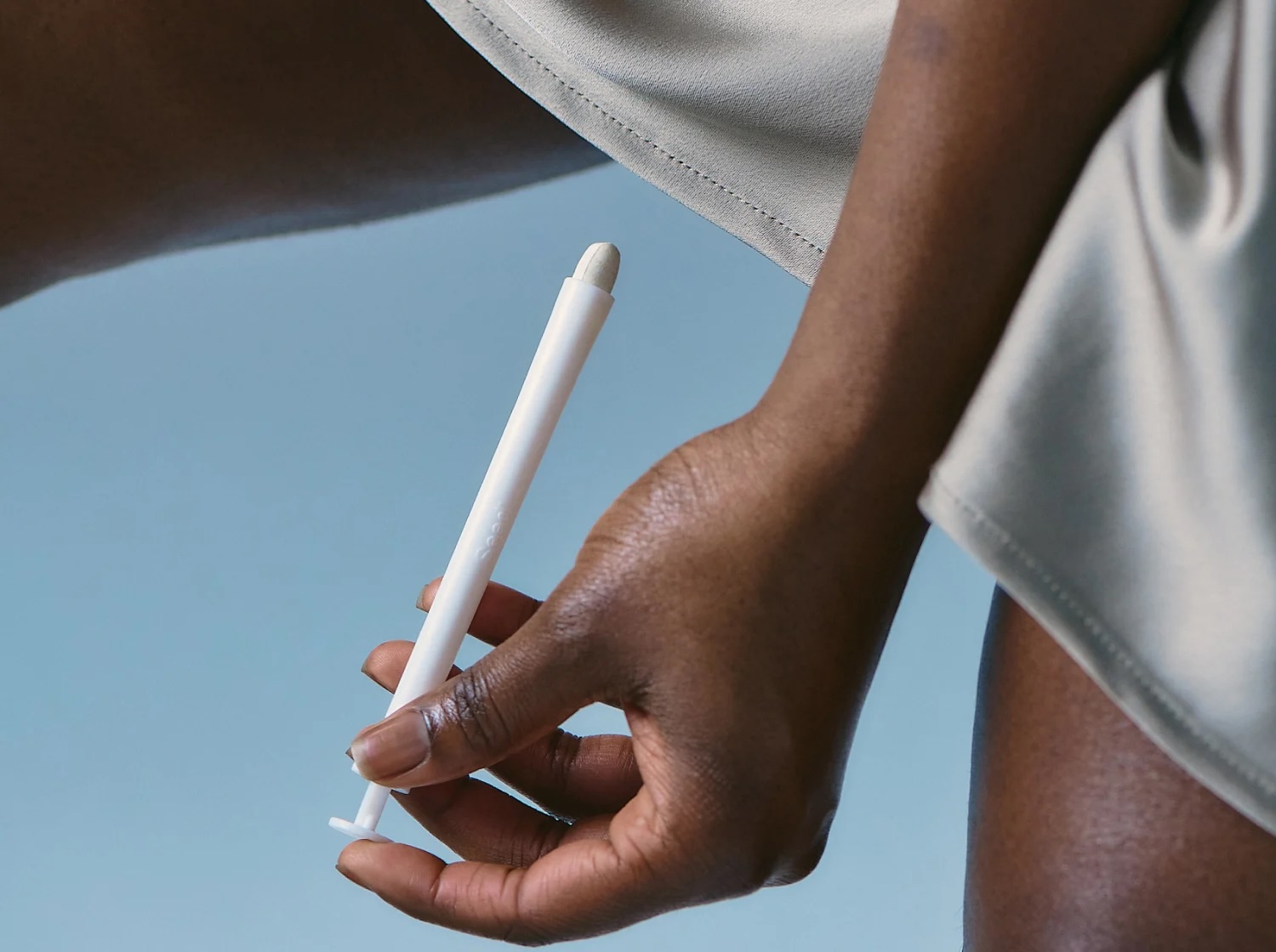 A person holds a white applicator-style device near their upper thigh, with a light blue background and neutral clothing visible.