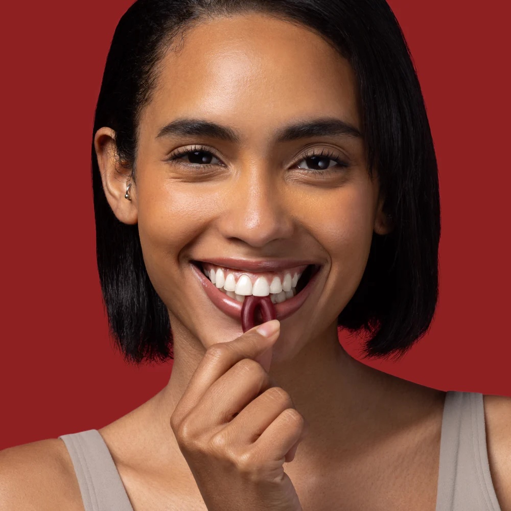 Woman with short dark hair smiles while holding a red capsule-shaped supplement near her mouth against a solid red background.