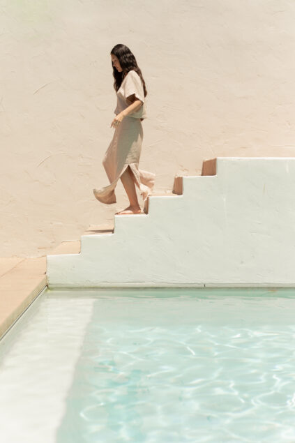 A woman in a light dress walks down white stairs beside a pool with clear water, against a beige textured wall.