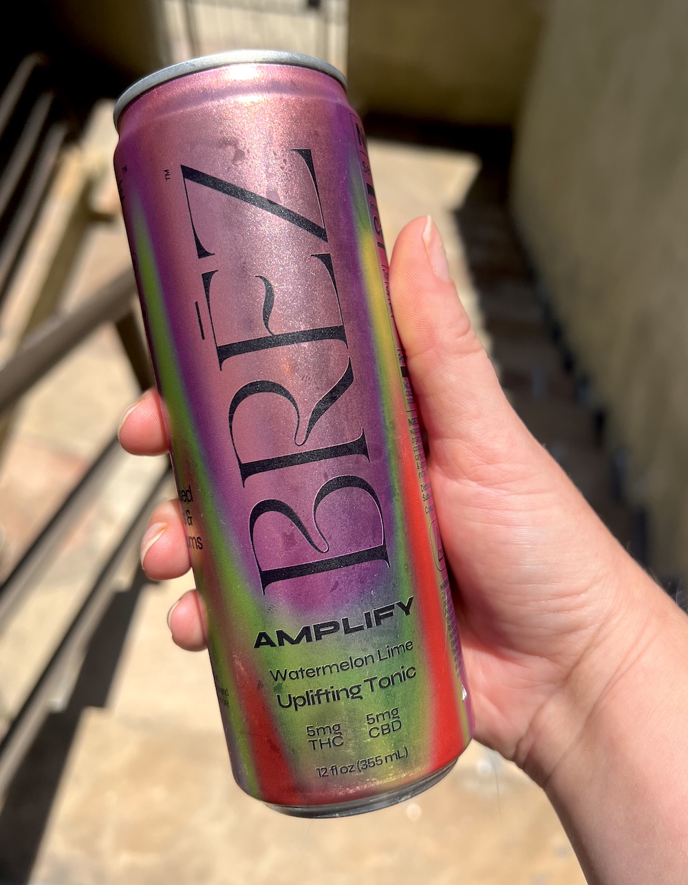 A hand holds a can of BRĒZ Amplify Watermelon Lime Uplifting Tonic, containing 5mg THC and 5mg CBD, against an outdoor background.