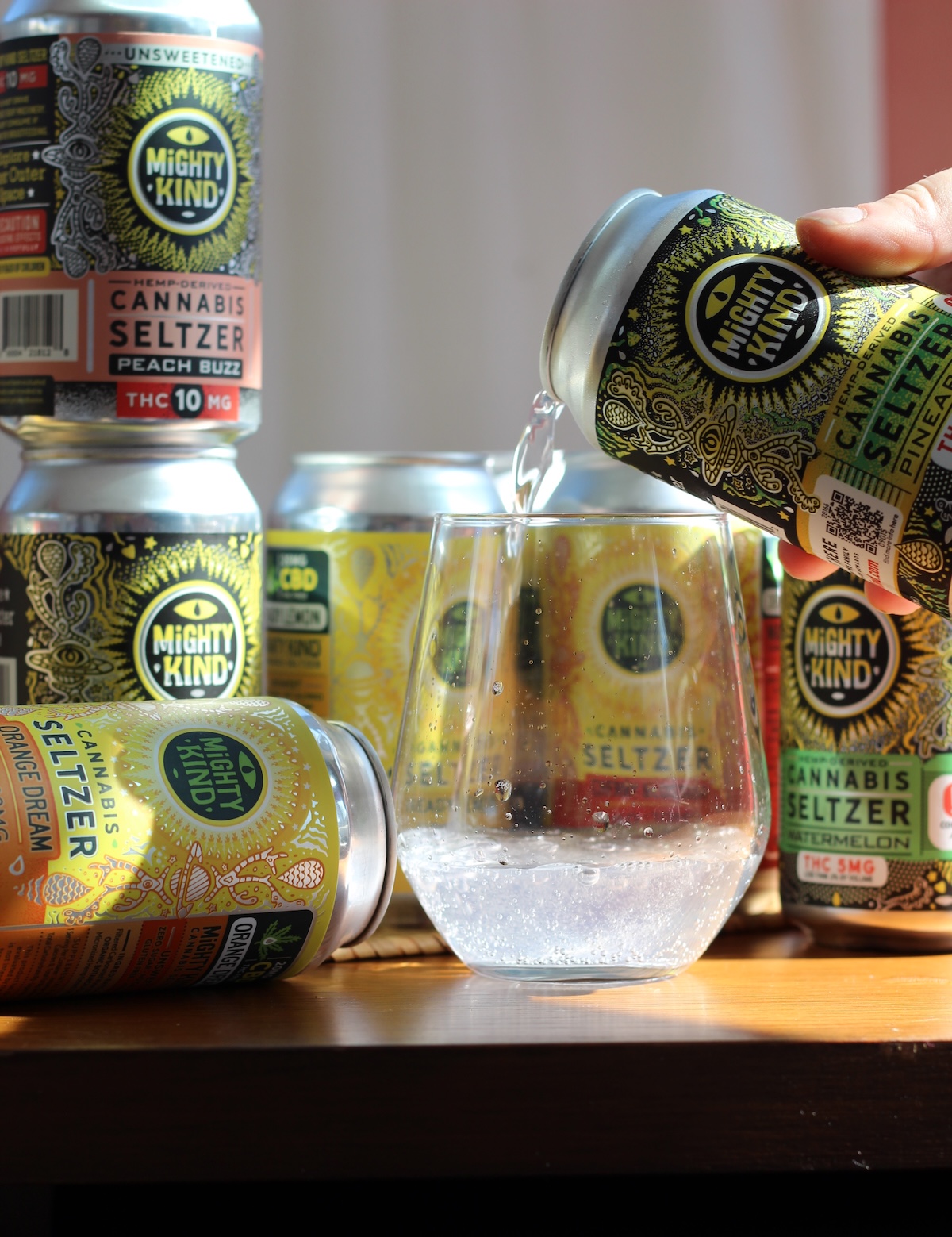 A hand pours Mighty Kind cannabis seltzer into a glass, with several cans of different flavors displayed on a wooden surface in sunlight.