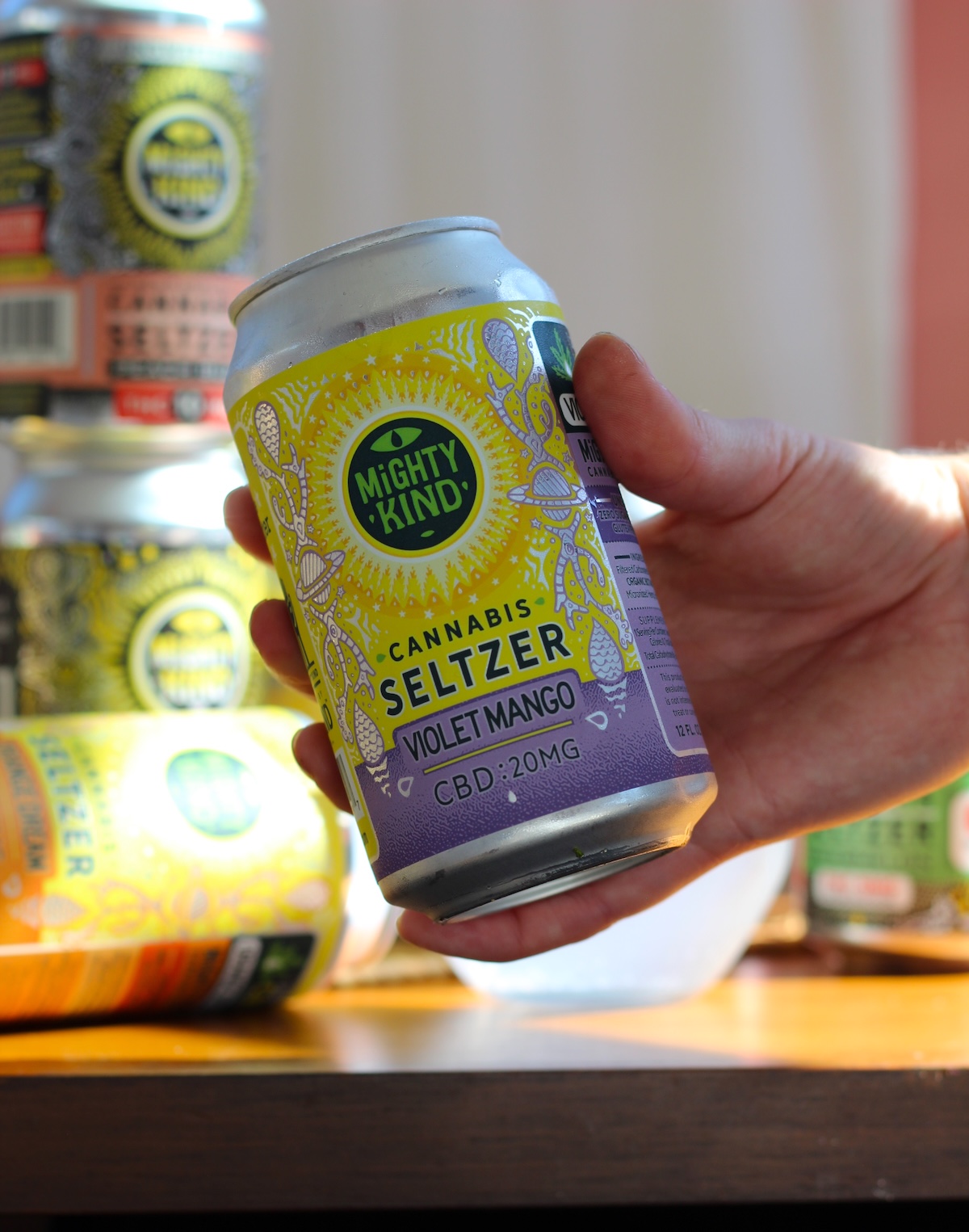 A hand holding a can of Mighty Kind Cannabis Seltzer, Violet Mango flavor, labeled with 20mg CBD. Other cans are visible in the background on a wooden surface.