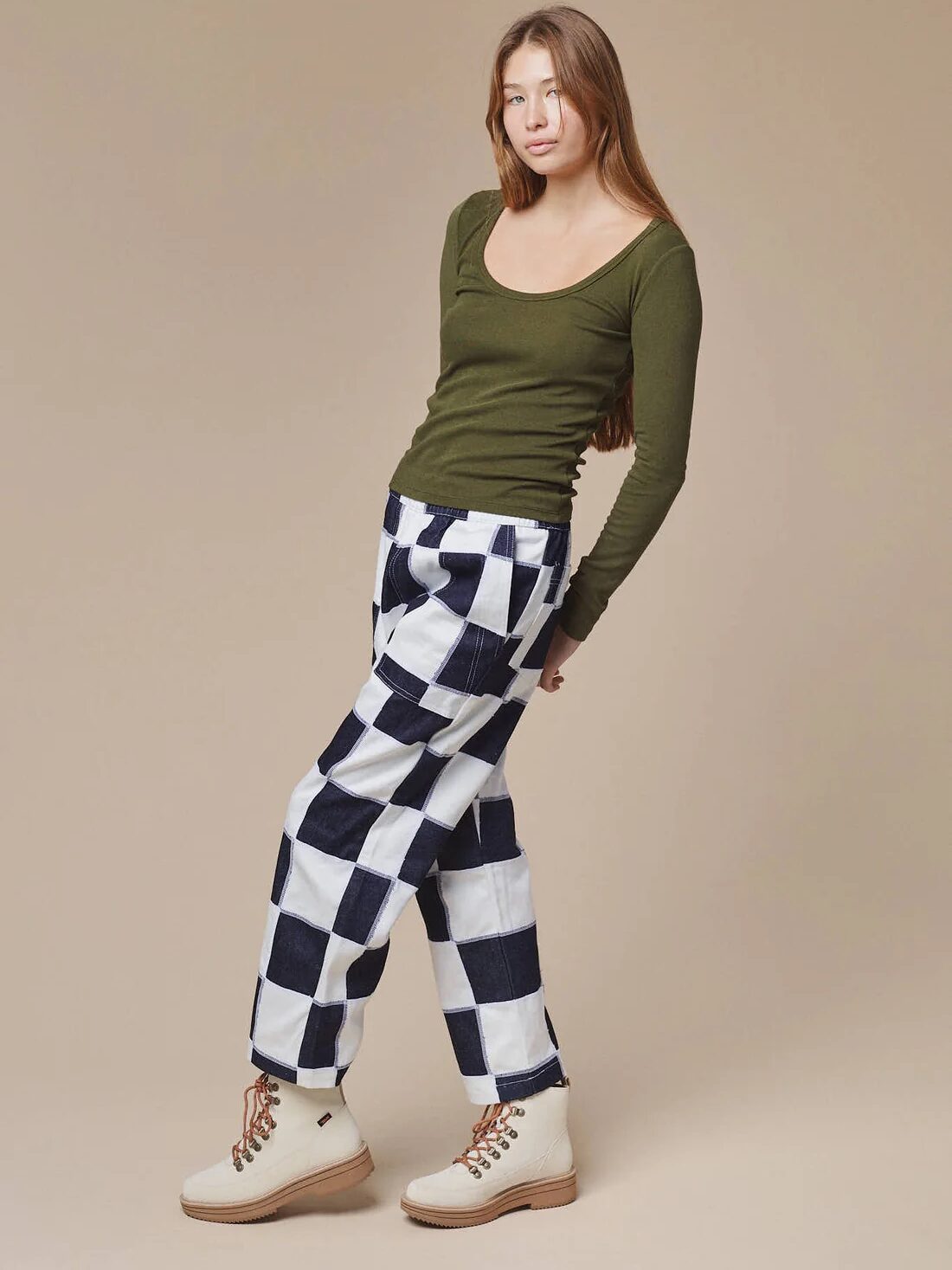 A person stands against a beige background, wearing an olive green long-sleeve top, black and white checkered pants, and beige boots.