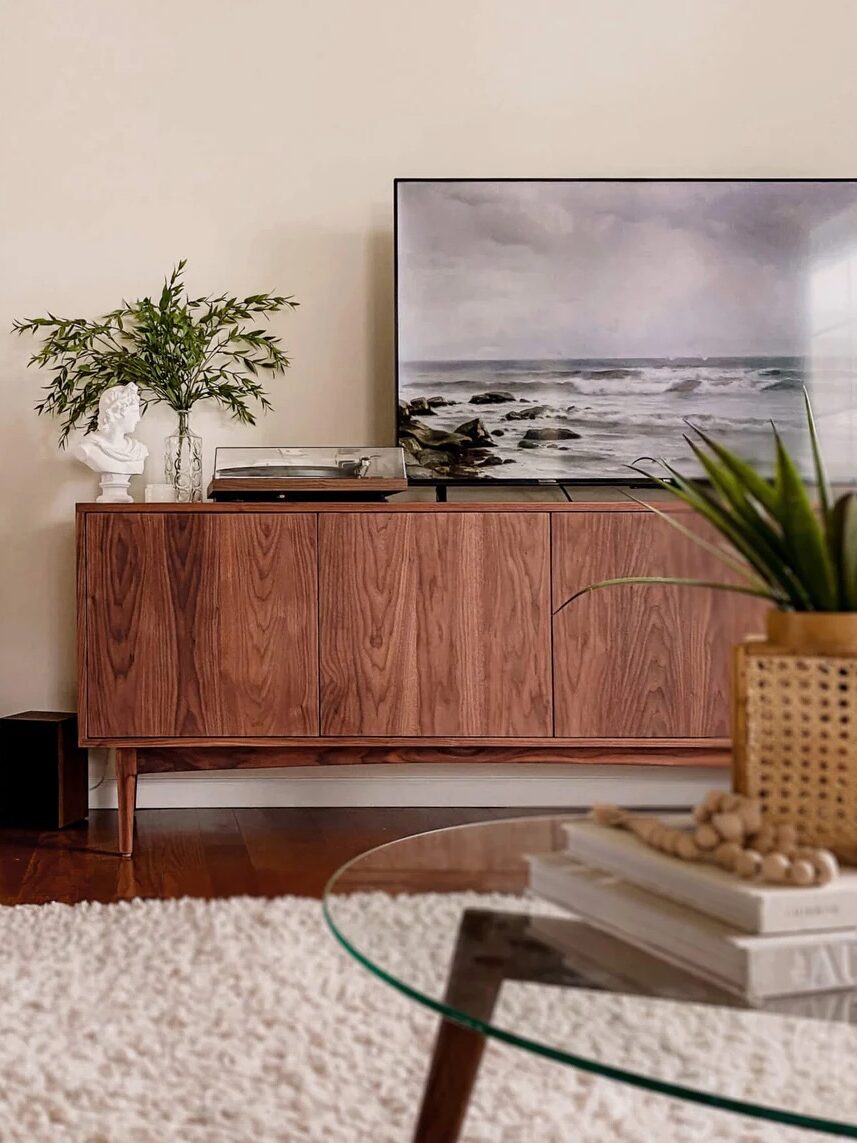 A modern living room with a wooden sideboard, flatscreen TV, potted plants, a large mirror, glass coffee table, and a textured white rug on a wooden floor.