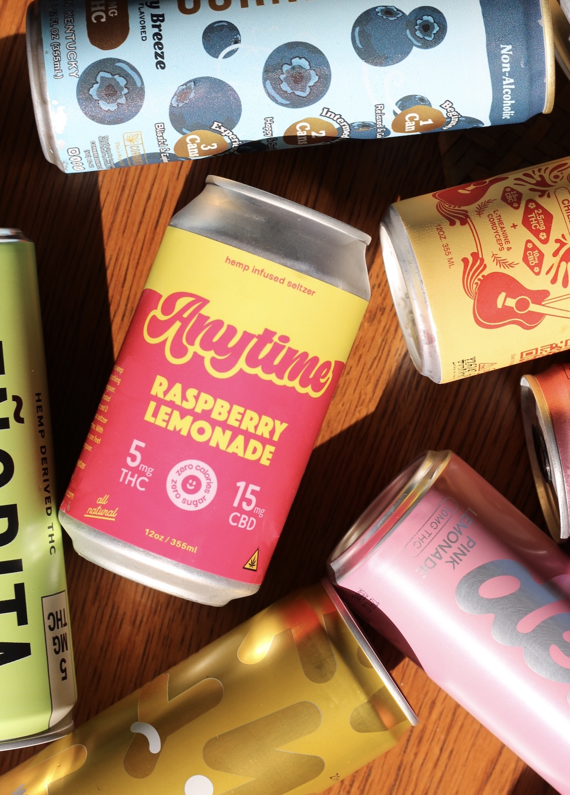 Several colorful beverage cans, including a prominently placed "Anytime Raspberry Lemonade" hemp-infused seltzer, are arranged on a wooden surface.