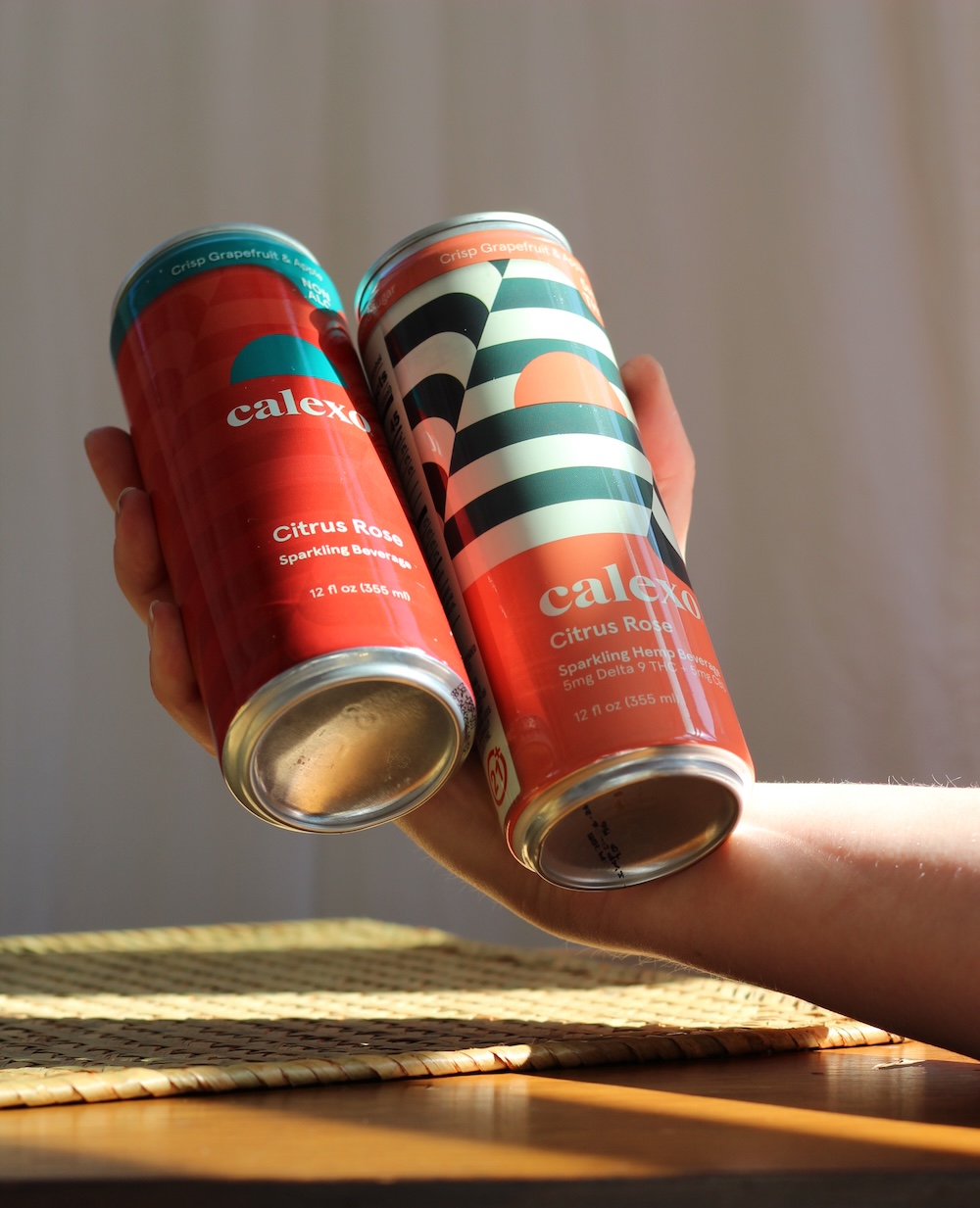 A hand holds two cans of Calexo Citrus Rose sparkling hemp beverage, featuring colorful geometric designs, against a softly lit background.