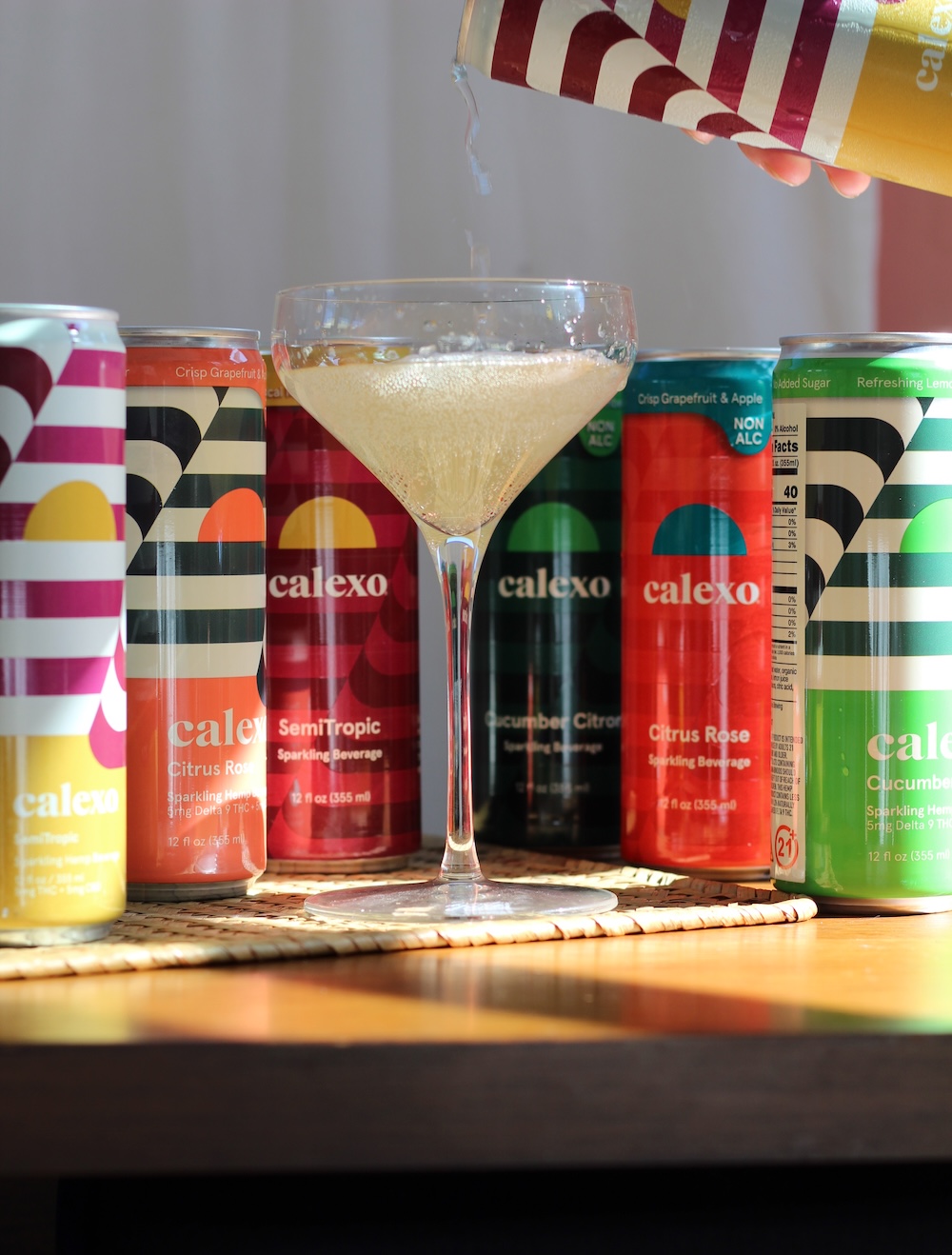 A glass is being filled with a fizzy drink poured from a striped can, with several colorful Calexo cans displayed in the background.