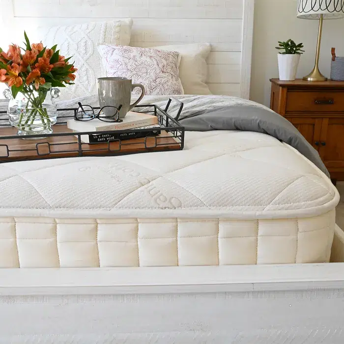 A neatly made bed with a white mattress, grey comforter, and a wooden tray holding flowers, a mug, glasses, and a book. A nightstand with a lamp is beside the bed.