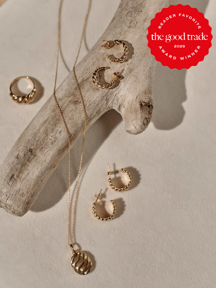 Gold jewelry including hoop earrings, rings, and a pendant necklace displayed on a piece of driftwood, with a red "The Good Trade 2025 Award Winner" badge in the corner.