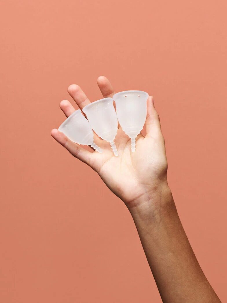 A hand holding three transparent menstrual cups against a solid peach-colored background.