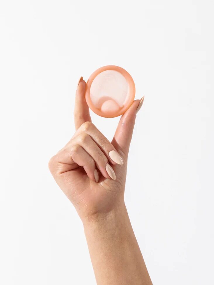 A hand with manicured nails holds up a pink contraceptive diaphragm against a plain white background.