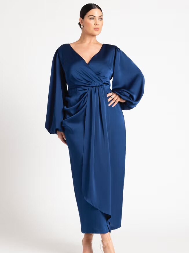 A woman poses in a deep blue, long-sleeve, V-neck satin dress with a draped front detail and midi length, wearing nude heels against a plain white background.