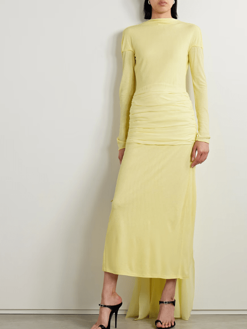 A person wearing a long-sleeved, pale yellow maxi dress with ruched detailing at the waist and black high-heeled sandals, standing against a plain light-colored wall.