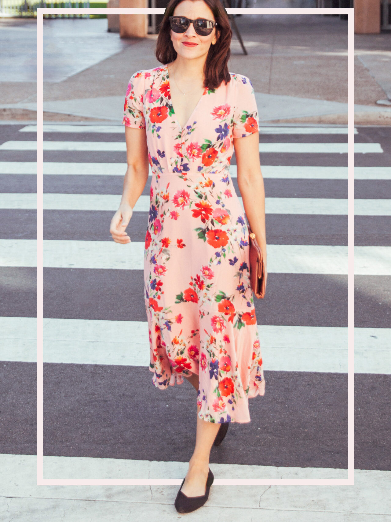 Woman wearing sunglasses and a pink floral dress stands on a city sidewalk with one leg slightly forward, holding a brown purse, as the sun casts long shadows.