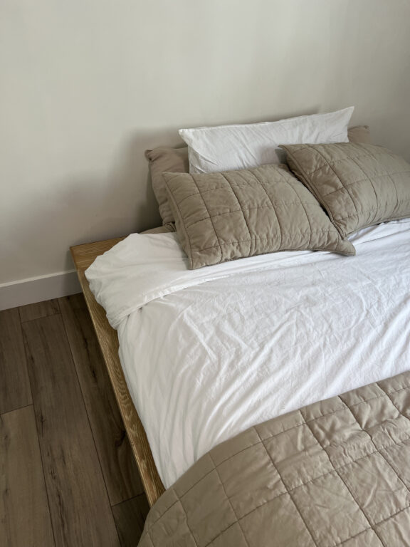 A neatly made bed with a white sheet, taupe quilted pillows, a matching comforter, and a wooden frame on a wooden floor against a plain white wall.