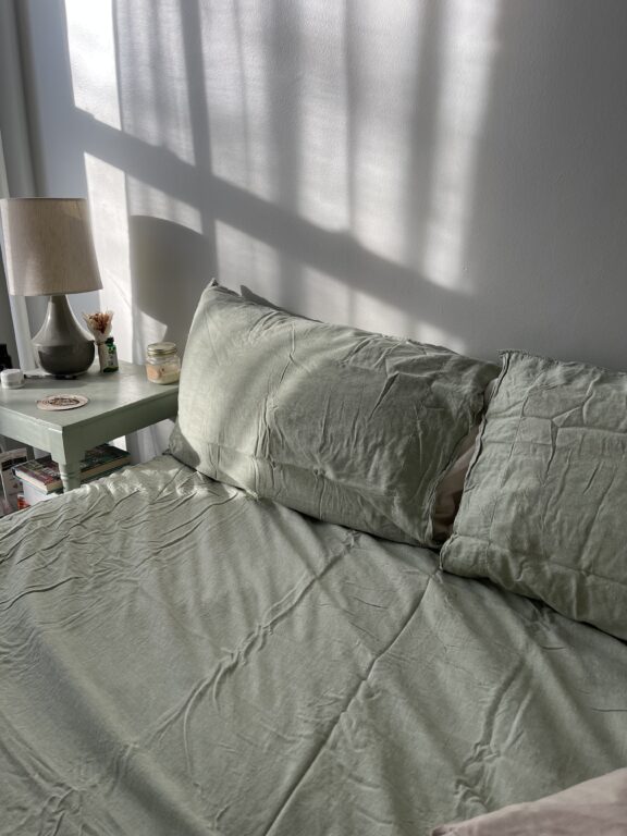 A neatly made bed with wrinkled light green sheets sits next to a small green nightstand with a lamp, candle, and a small plant; sunlight and shadows fall across the scene.