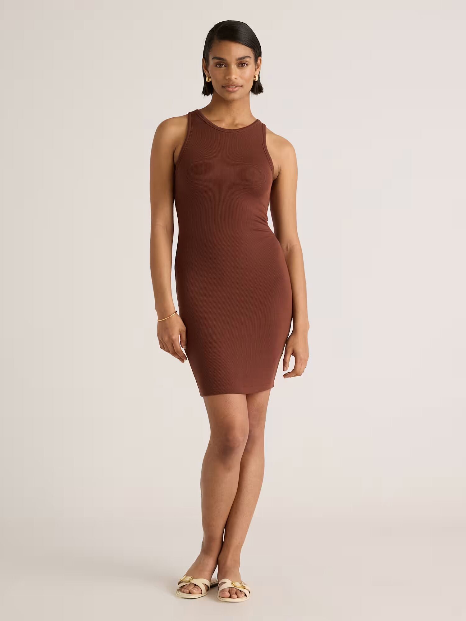 A woman wearing a sleeveless, fitted brown dress and sandals stands against a plain background, facing forward with a neutral expression.