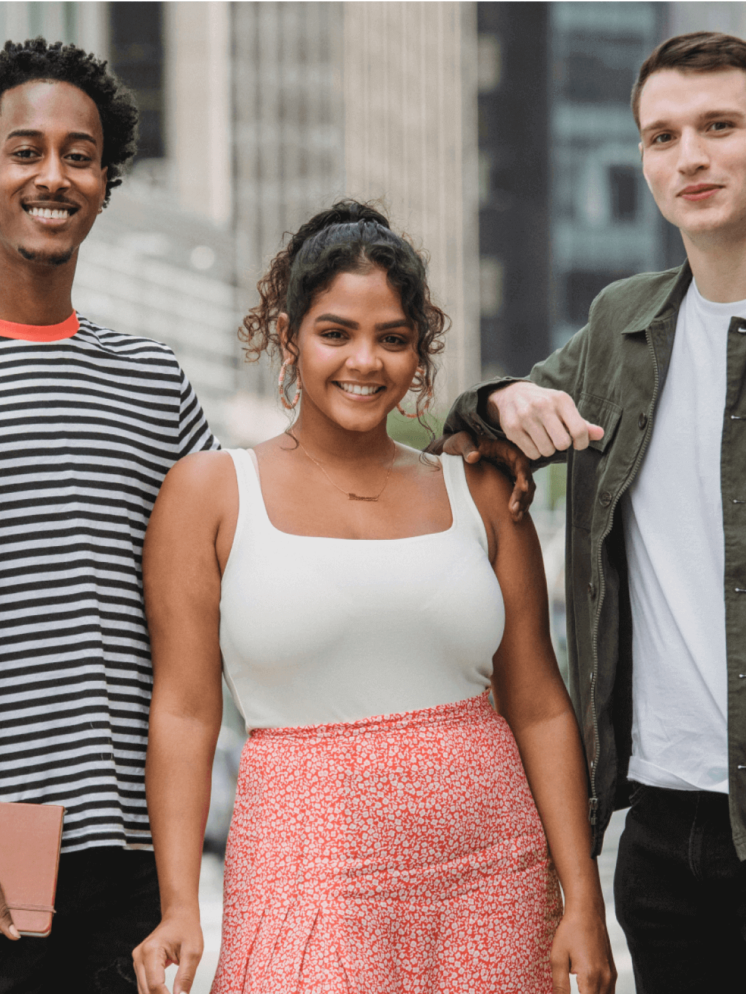 Three people stand close together outdoors in a city setting, smiling at the camera. One holds a notebook, and tall buildings are visible in the background.