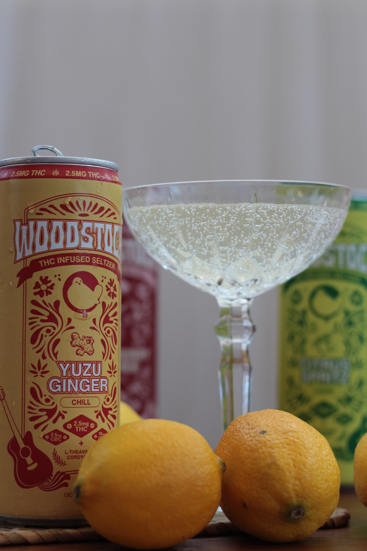 A yellow can of Woodstock yuzu ginger THC-infused seltzer stands next to a coupe glass filled with a sparkling drink and three whole lemons, with two other cans blurred in the background.