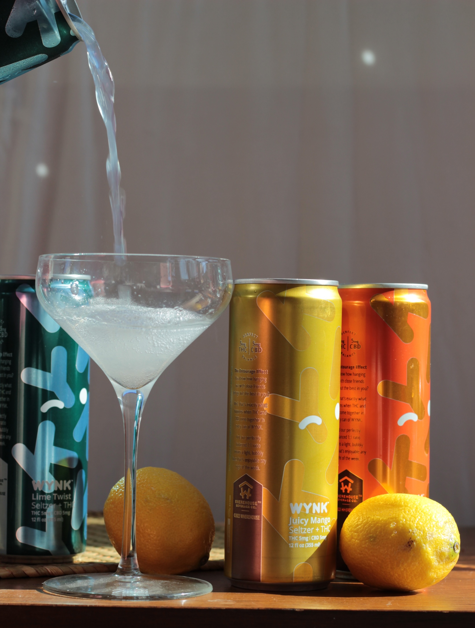A clear liquid is being poured into a cocktail glass next to three WYNK canned drinks and two whole lemons on a wooden surface.