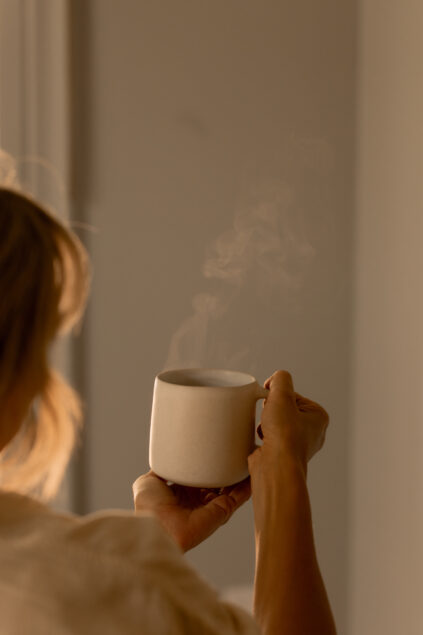 A person holding a white ceramic mug with steam rising, viewed from behind against a neutral background.