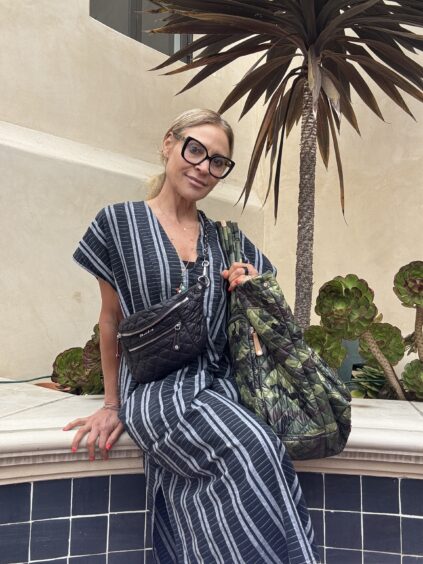 A woman in a striped dress sits on a tiled ledge, holding two quilted bags, with succulents and a tall plant in the background.