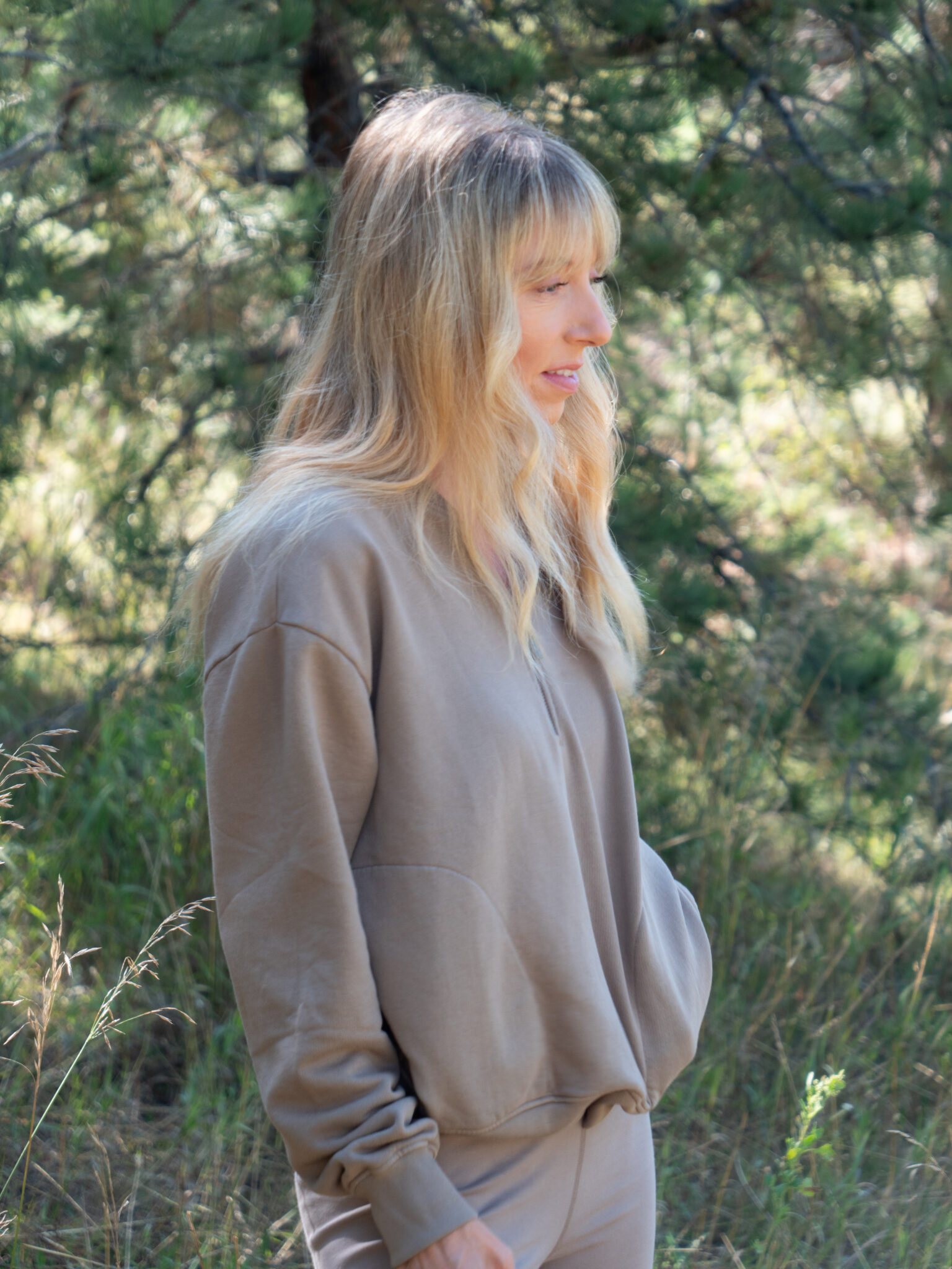 A woman with long blonde hair wearing a tan sweatshirt stands outdoors in a grassy, wooded area.