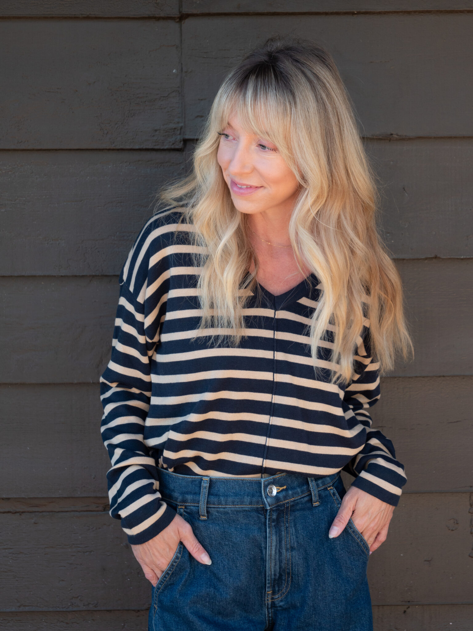 A woman with long blonde hair wearing a black and beige striped top and blue jeans stands in front of a dark wooden wall with her hands in her pockets.