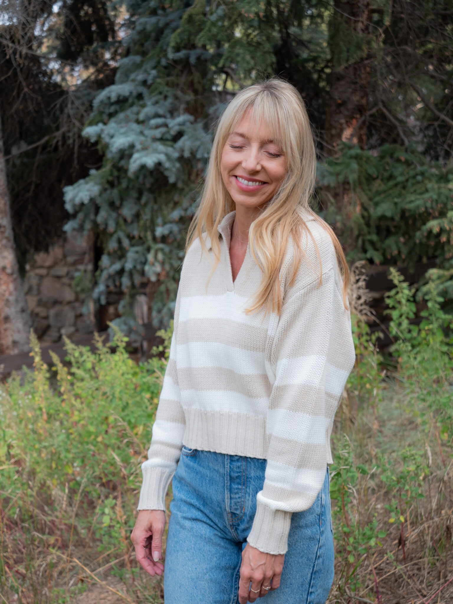 A woman with long blonde hair wears a cream striped sweater and blue jeans, standing outdoors in front of green foliage and trees, smiling with her eyes closed.