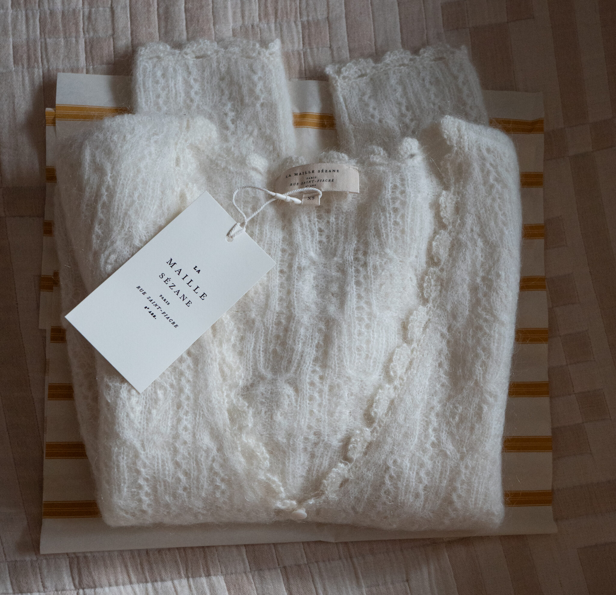 A folded white knitted sweater with tags attached, placed on striped tissue paper, lying on a textured surface.