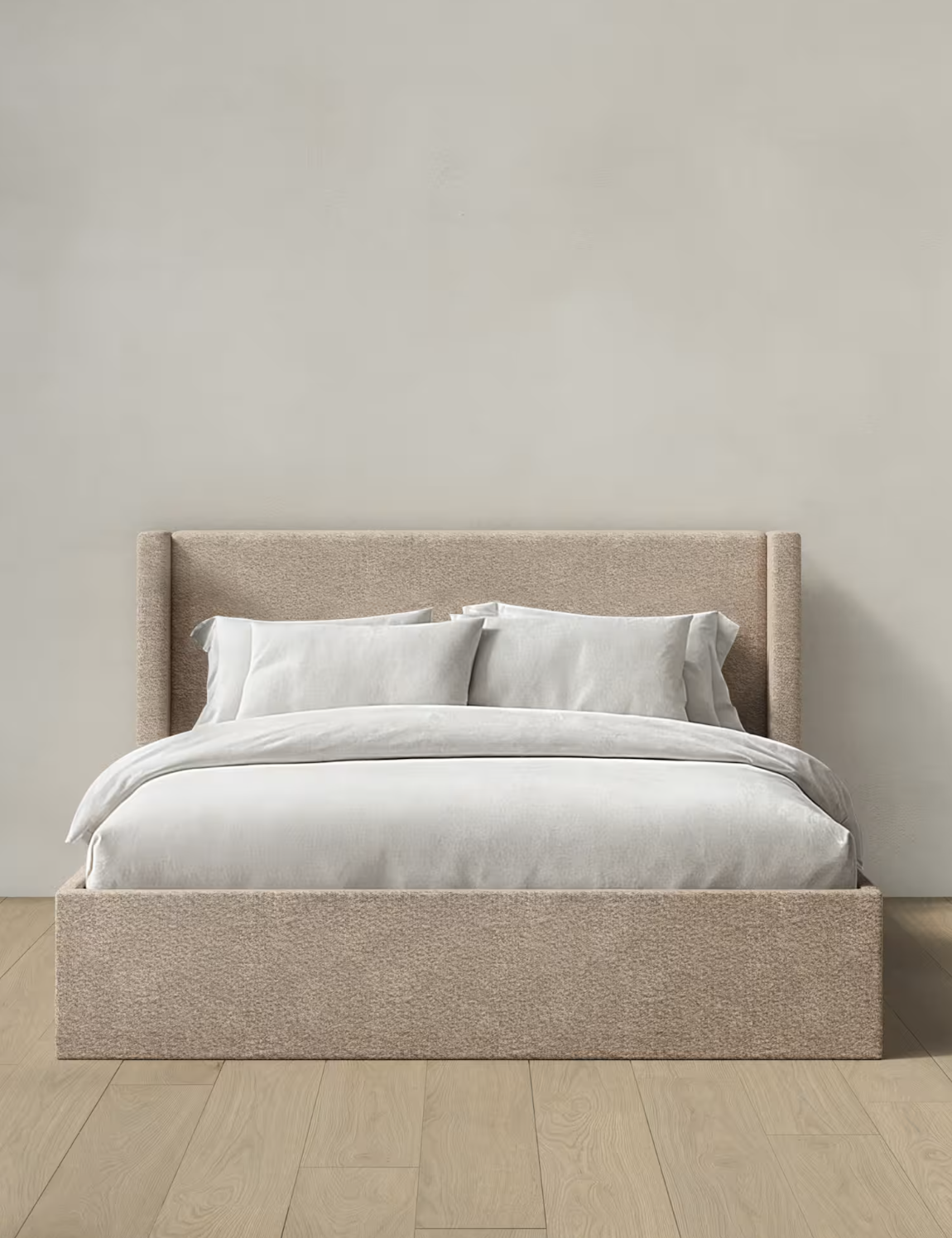 A beige upholstered bed with white bedding and pillows, set against a plain light-colored wall on a wooden floor.